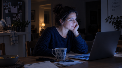 A woman sits alone at a kitchen table late at night, lit by an open laptop screen, the rest of the room dark.